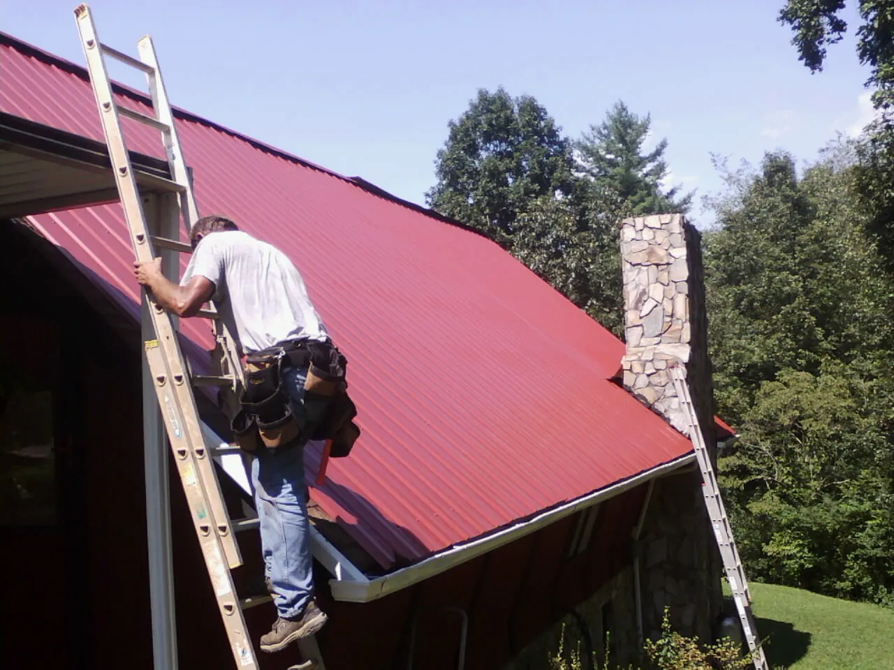 Roofer on a ladder beside a red metal roof and stone chimney — Soffit & Fascia Repair by Roofing Brothers in Chester